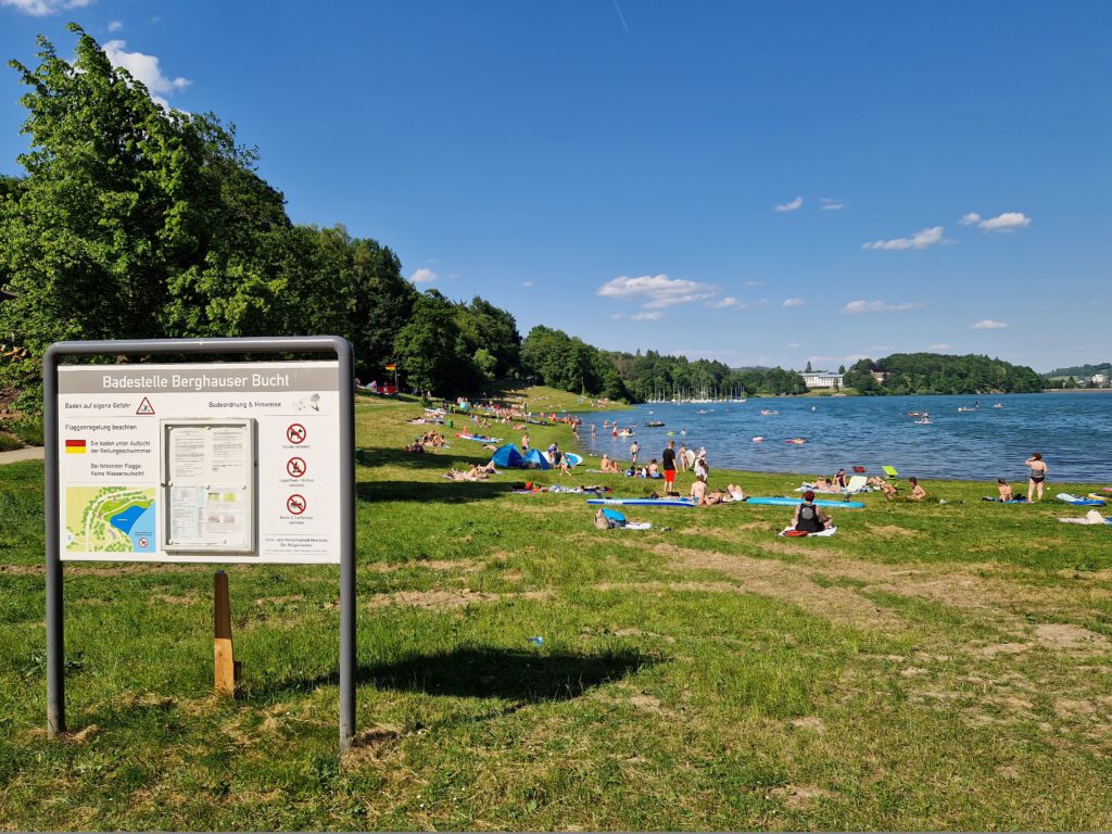 Ein Blick über die Berghauser Bucht, die größte Badestelle am Hennesee. Im Vordergrund ist eine Liegewiese, im Hintergrund des Bildes ist der Hennesee zu sehen.